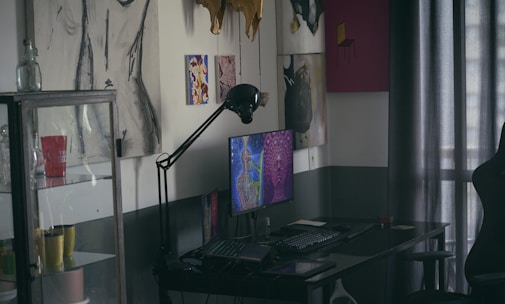 A dimly lit room features a desk with a computer displaying colorful artwork on the screen. On the wall are various art pieces, including abstract and figure drawings. A black desk lamp arches over the computer. To the left, a glass cabinet holds multiple mugs, one of which is red with white text. Curtains filter light in the background.