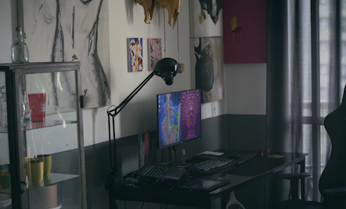 A dimly lit room features a desk with a computer displaying colorful artwork on the screen. On the wall are various art pieces, including abstract and figure drawings. A black desk lamp arches over the computer. To the left, a glass cabinet holds multiple mugs, one of which is red with white text. Curtains filter light in the background.