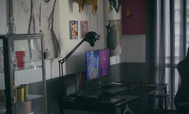 A dimly lit room features a desk with a computer displaying colorful artwork on the screen. On the wall are various art pieces, including abstract and figure drawings. A black desk lamp arches over the computer. To the left, a glass cabinet holds multiple mugs, one of which is red with white text. Curtains filter light in the background.