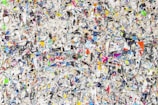 A chaotic and colorful mixture of torn paper pieces with various text, images, and colors scattered densely throughout. The image is a collage that creates a textured and vibrant appearance, making it difficult to focus on any single piece. The torn papers overlap and intersect, offering a visual representation of randomness and disorder.