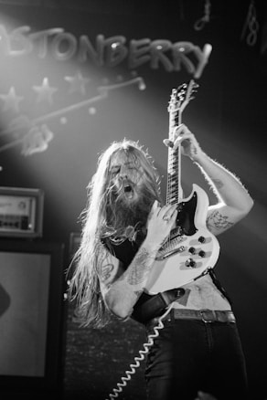 A musician with long hair energetically playing an electric guitar on stage under dim lighting. They are surrounded by amplifiers and stage equipment, with a sign in the background that reads 'STONBERRY.' The atmosphere suggests a live rock performance.