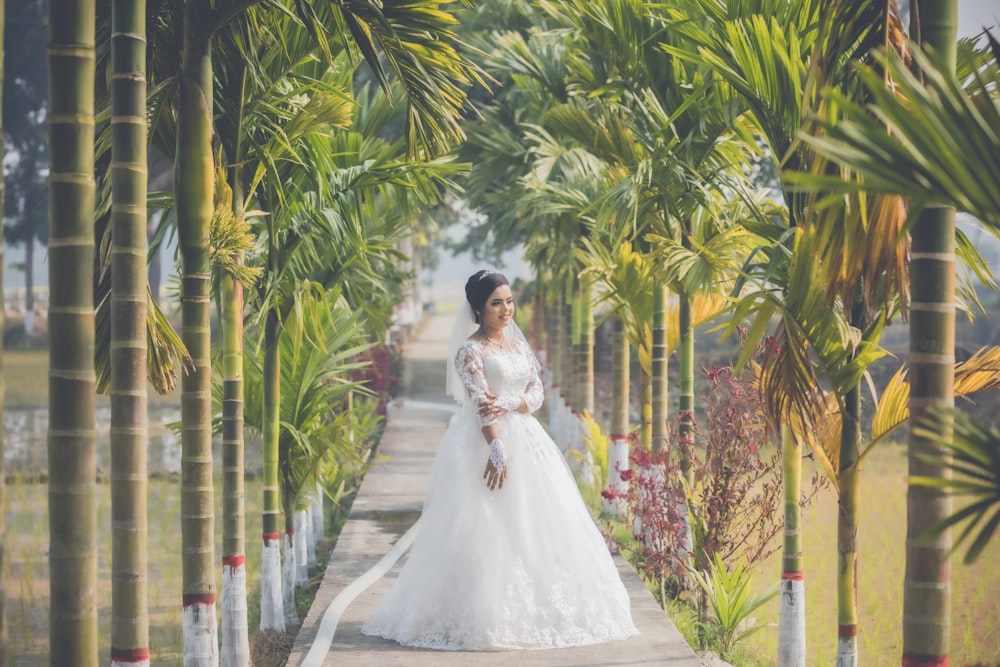 donna in abito da sposa bianco che cammina sul sentiero tra le palme durante il giorno