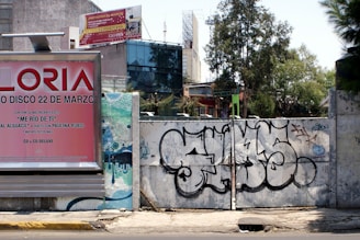 A street scene featuring a large billboard advertising a music album, positioned next to a graffitied metal gate. The billboard has a pink and red color scheme with text promoting an album and a song by a specific artist. Behind the gate, there are trees and urban structures, including a variety of commercial and residential buildings.