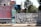 A street scene featuring a large billboard advertising a music album, positioned next to a graffitied metal gate. The billboard has a pink and red color scheme with text promoting an album and a song by a specific artist. Behind the gate, there are trees and urban structures, including a variety of commercial and residential buildings.