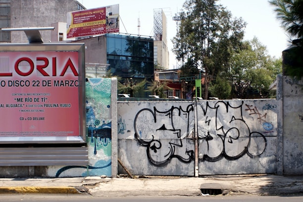 A street scene featuring a large billboard advertising a music album, positioned next to a graffitied metal gate. The billboard has a pink and red color scheme with text promoting an album and a song by a specific artist. Behind the gate, there are trees and urban structures, including a variety of commercial and residential buildings.
