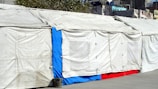 A long makeshift tent is set up outdoors with beige tarps forming the walls and roof. A section of the tent features a bright blue and red panel towards the bottom. The tarps are slightly wrinkled and secured with ties, with a trash can placed on the ground nearby. Urban buildings and foliage are visible in the background.