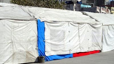 A long makeshift tent is set up outdoors with beige tarps forming the walls and roof. A section of the tent features a bright blue and red panel towards the bottom. The tarps are slightly wrinkled and secured with ties, with a trash can placed on the ground nearby. Urban buildings and foliage are visible in the background.