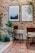A cozy interior setup features a brick wall adorned with two framed pictures, one showcasing a misty forest and the other with an inspirational quote. Several potted plants add a touch of greenery, and wooden chairs are placed around a tiled floor with intricate patterns.