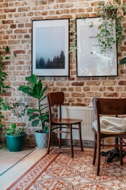 A cozy interior setup features a brick wall adorned with two framed pictures, one showcasing a misty forest and the other with an inspirational quote. Several potted plants add a touch of greenery, and wooden chairs are placed around a tiled floor with intricate patterns.