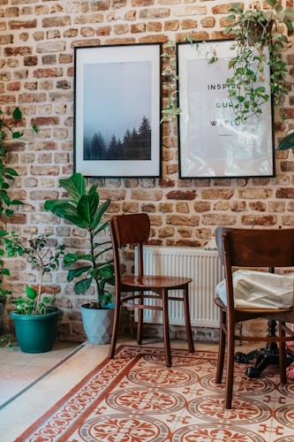 A cozy interior setup features a brick wall adorned with two framed pictures, one showcasing a misty forest and the other with an inspirational quote. Several potted plants add a touch of greenery, and wooden chairs are placed around a tiled floor with intricate patterns.