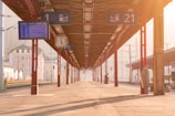 A train platform bathed in warm sunlight features a long roof supported by red columns. Overhead signs indicate platform numbers and departure details. A large overhead clock is visible alongside an electronic timetable display.