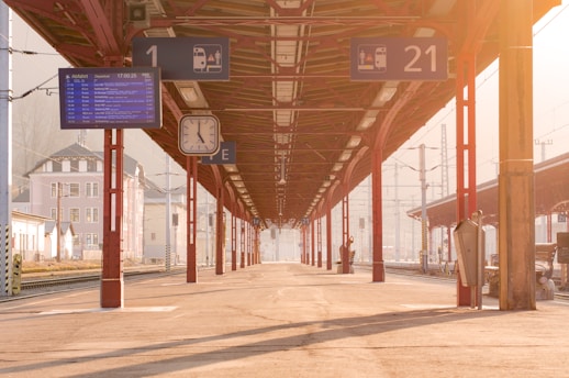 A train platform bathed in warm sunlight features a long roof supported by red columns. Overhead signs indicate platform numbers and departure details. A large overhead clock is visible alongside an electronic timetable display.