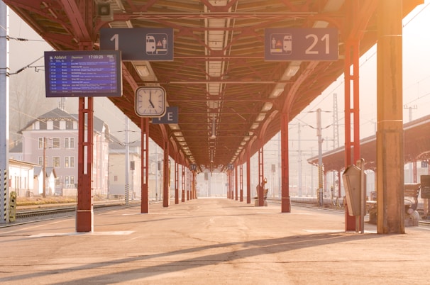 A train platform bathed in warm sunlight features a long roof supported by red columns. Overhead signs indicate platform numbers and departure details. A large overhead clock is visible alongside an electronic timetable display.