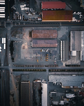 An aerial view of an industrial area with multiple large buildings and warehouses surrounded by roads. Vehicles and construction equipment are visible within the yard. The layout is grid-like, with intersections and train tracks appearing alongside the buildings.