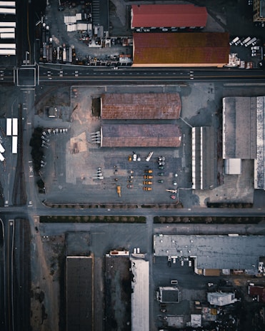 An aerial view of an industrial area with multiple large buildings and warehouses surrounded by roads. Vehicles and construction equipment are visible within the yard. The layout is grid-like, with intersections and train tracks appearing alongside the buildings.