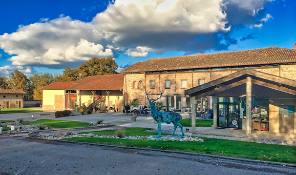 A scenic view featuring a rustic building with brick walls and a tiled roof, surrounded by a large courtyard. In the foreground, a life-sized statue of a blue stag stands on a patch of well-manicured grass. The sky above is vivid blue with fluffy white clouds casting shadows on the landscape.
