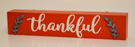 A rectangular wooden sign with an orange background featuring the word 'thankful' in elegant white script. Two metallic leaf embellishments are positioned on each side of the word.