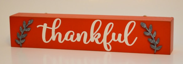 A rectangular wooden sign with an orange background featuring the word 'thankful' in elegant white script. Two metallic leaf embellishments are positioned on each side of the word.