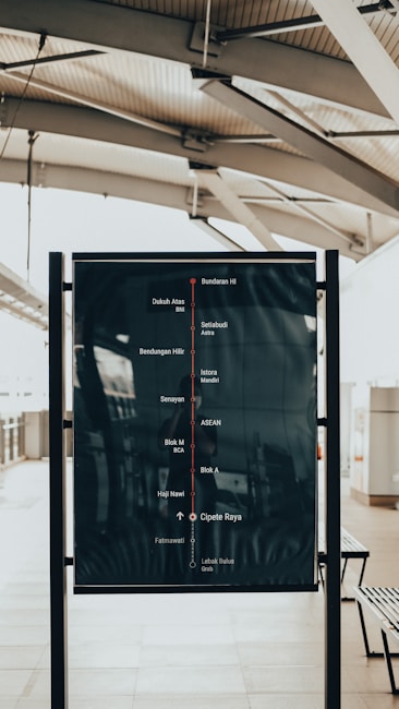 A public transportation map displayed on a black signboard, featuring station names and lines in a railway terminal. The background includes a modern architectural ceiling with metallic beams and a tiled floor.