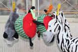 Several stuffed animal toys hang from a rope secured with colorful clothespins. The toys include a plush raccoon, a toy that resembles a slice of watermelon, and a zebra head with stripes. The background is a wall with metal bars, possibly a window frame.