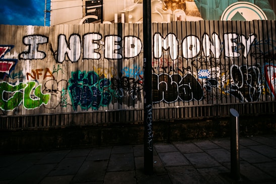 A corrugated metal fence is covered in graffiti, with the most prominent words being 'I NEED MONEY' in bold white letters outlined in black. Various colorful tags and messages in shades of green, blue, and black surround the main text. A dark vertical pole divides the image in the center, and a small blue and white sign is visible on the right.