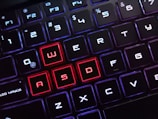 The image displays a close-up view of a keyboard with backlit keys. Most of the keys are illuminated in white, while the keys W, A, S, and D are highlighted in red, drawing attention to them. The arrangement suggests a focus on gaming-related keyboard use, as these keys are often used in games for navigation.