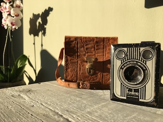 A vintage camera with the label 'Fodor-Box' sits on a rustic wooden surface next to a brown leather bag. To the left, vibrant orchids cast a dramatic shadow against a light green wall, creating a nostalgic and serene ambiance.