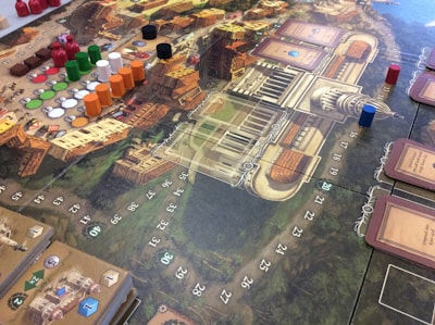 A detailed view of a board game setup with a complex game board featuring a map-like design. Various colored game pieces, including small discs and cylindrical markers in red, green, black, white, and orange, are arranged across the board. There are also decks of cards on the board, each having illustrations and text. The board contains numbered spaces and intricate graphics representing a European city or town. The game appears to be a strategic or historical themed game.