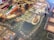 A detailed view of a board game setup with a complex game board featuring a map-like design. Various colored game pieces, including small discs and cylindrical markers in red, green, black, white, and orange, are arranged across the board. There are also decks of cards on the board, each having illustrations and text. The board contains numbered spaces and intricate graphics representing a European city or town. The game appears to be a strategic or historical themed game.