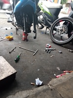 A motorcycle repair shop scene with a bike elevated on a stand, various tools such as wrenches and screwdrivers scattered on the ground, along with trash and debris. A detached wheel is visible, indicating maintenance work is in progress.