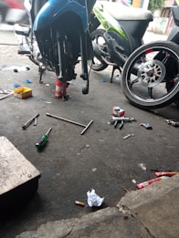 A motorcycle repair shop scene with a bike elevated on a stand, various tools such as wrenches and screwdrivers scattered on the ground, along with trash and debris. A detached wheel is visible, indicating maintenance work is in progress.