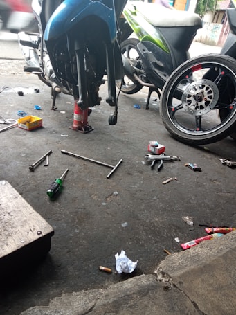 A motorcycle repair shop scene with a bike elevated on a stand, various tools such as wrenches and screwdrivers scattered on the ground, along with trash and debris. A detached wheel is visible, indicating maintenance work is in progress.