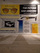 A mural painted on a brick wall depicts various illustrations and texts. A pair of sunglasses is shown in two variations, with 'FLOWS' written above one of them. There is bold text stating 'THE FUTURE HAS ARRIVED'. Another section features an ear with sound waves, and a text promoting 'Immersive High-Quality Listening'. The hashtag '#GETINFLOWS' is also present.