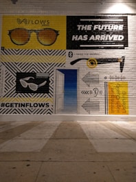 A mural painted on a brick wall depicts various illustrations and texts. A pair of sunglasses is shown in two variations, with 'FLOWS' written above one of them. There is bold text stating 'THE FUTURE HAS ARRIVED'. Another section features an ear with sound waves, and a text promoting 'Immersive High-Quality Listening'. The hashtag '#GETINFLOWS' is also present.