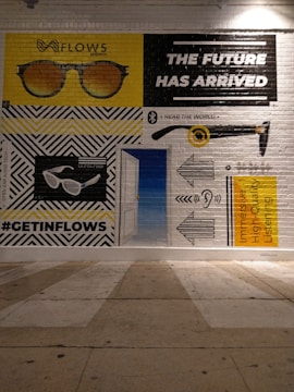 A mural painted on a brick wall depicts various illustrations and texts. A pair of sunglasses is shown in two variations, with 'FLOWS' written above one of them. There is bold text stating 'THE FUTURE HAS ARRIVED'. Another section features an ear with sound waves, and a text promoting 'Immersive High-Quality Listening'. The hashtag '#GETINFLOWS' is also present.
