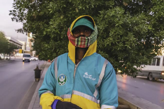 A person is wearing a bright blue jacket with yellow and gray accents, featuring Arabic writing and a logo. They are also wearing a plaid scarf and a cap with the hood of the jacket pulled up. The background includes a street with some vehicles and a tree, suggesting an urban setting.