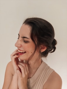A person with dark hair is smiling warmly, looking relaxed and content. They are wearing a sleeveless beige top and their hair is tied back. Their expression conveys joy and friendliness.