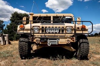 A rugged military-style vehicle with large tires and a beige exterior is parked on a grassy field. The sky is clear with some clouds, and there are trees visible in the background. The vehicle has a prominent front grill and license plate, suggesting it is designed for off-road or all-terrain use.