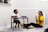 A young boy and a woman are engaged in an activity together. The boy is sitting at a small table drawing or writing, while the woman is sitting on the floor, appearing to be encouraging or helping him. There are some colorful papers on the floor beside them.