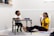 A young boy and a woman are engaged in an activity together. The boy is sitting at a small table drawing or writing, while the woman is sitting on the floor, appearing to be encouraging or helping him. There are some colorful papers on the floor beside them.