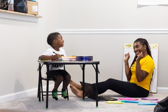 A young boy and a woman are engaged in an activity together. The boy is sitting at a small table drawing or writing, while the woman is sitting on the floor, appearing to be encouraging or helping him. There are some colorful papers on the floor beside them.
