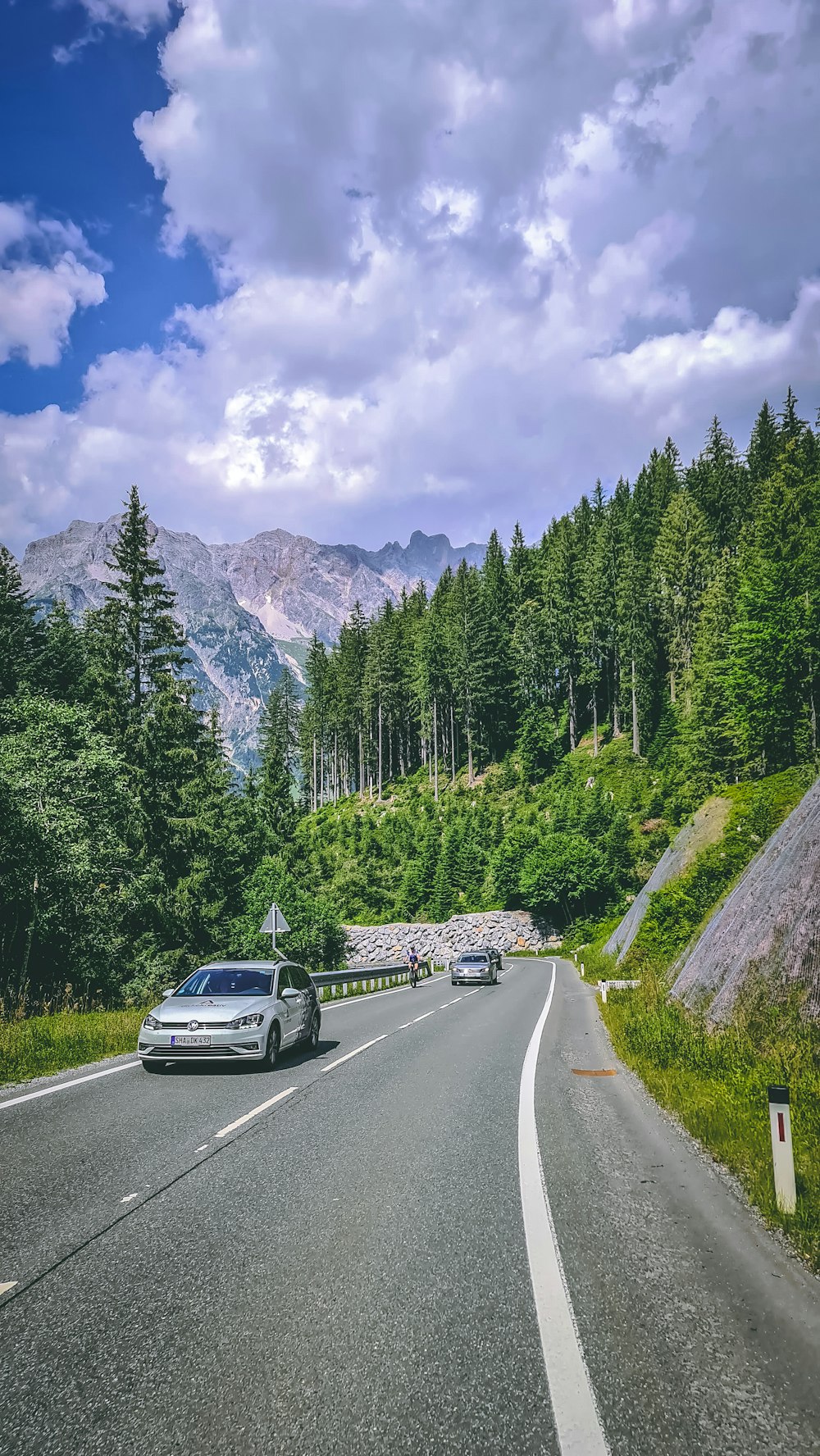 black car on road near green trees and mountain under blue sky and white clouds during