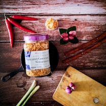 A jar of 'Bawang Goreng Sumenep' with a purple lid is positioned on a black pan, surrounded by various cooking ingredients. To the left are several red chili peppers and stalks of lemon grass. Near the top, there is a small bowl filled with crispy fried onions. On the right are cinnamon sticks, red shallots on green leaves, and a wooden cutting board with more shallots.