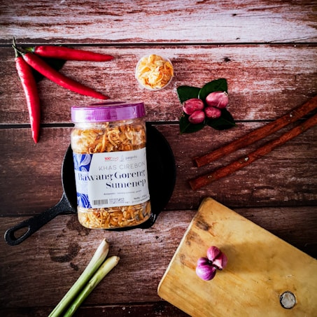 A jar of 'Bawang Goreng Sumenep' with a purple lid is positioned on a black pan, surrounded by various cooking ingredients. To the left are several red chili peppers and stalks of lemon grass. Near the top, there is a small bowl filled with crispy fried onions. On the right are cinnamon sticks, red shallots on green leaves, and a wooden cutting board with more shallots.