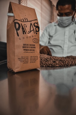 A brown paper bag of coffee labeled 'Café Pacas Gourmet' stands on a reflective surface. In the background, a person wearing a mask is inspecting a pile of roasted coffee beans.