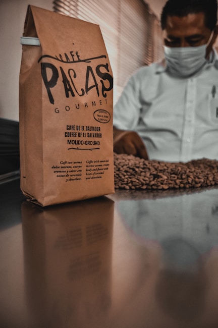 A brown paper bag of coffee labeled 'Caf&eacute; Pacas Gourmet' stands on a reflective surface. In the background, a person wearing a mask is inspecting a pile of roasted coffee beans.