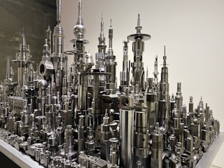 A collection of metal components and machine parts arranged vertically to resemble a futuristic cityscape. The elements include cylindrical and spherical shapes, gears, and various mechanical parts, all with a shiny metallic finish.