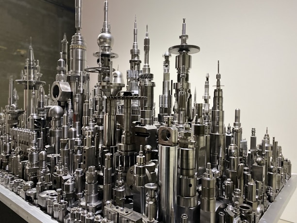 A collection of metal components and machine parts arranged vertically to resemble a futuristic cityscape. The elements include cylindrical and spherical shapes, gears, and various mechanical parts, all with a shiny metallic finish.