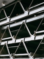 A close-up view of an industrial structure displaying several parallel metallic beams and cross-braces arranged in a geometric pattern. The beams are primarily composed of metal and exhibit a clean, angular design. A light fixture is visible at the top left corner.