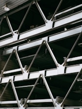A close-up view of an industrial structure displaying several parallel metallic beams and cross-braces arranged in a geometric pattern. The beams are primarily composed of metal and exhibit a clean, angular design. A light fixture is visible at the top left corner.